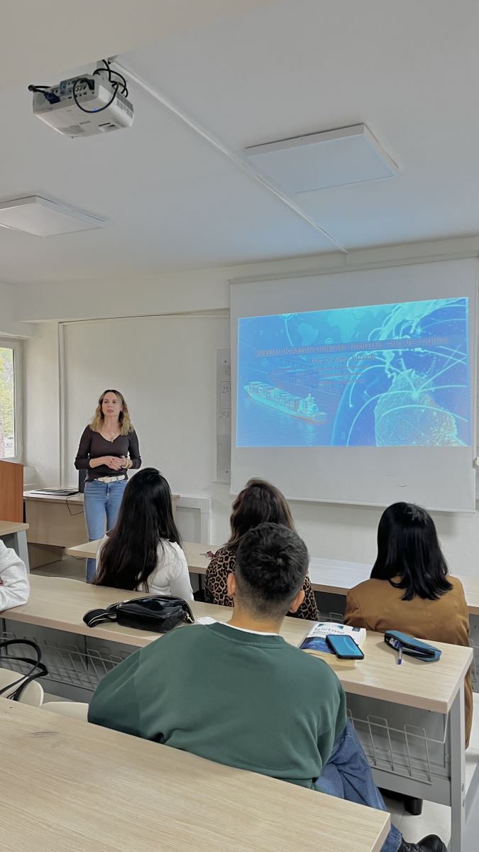 A woman is standing at the front of a classroom, giving a presentation. The projection behind her shows a background with a world map, a cargo ship, and digital lines. She is speaking to the students seated in rows, who are facing her and listening attentively. The room is bright and has a simple classroom layout.